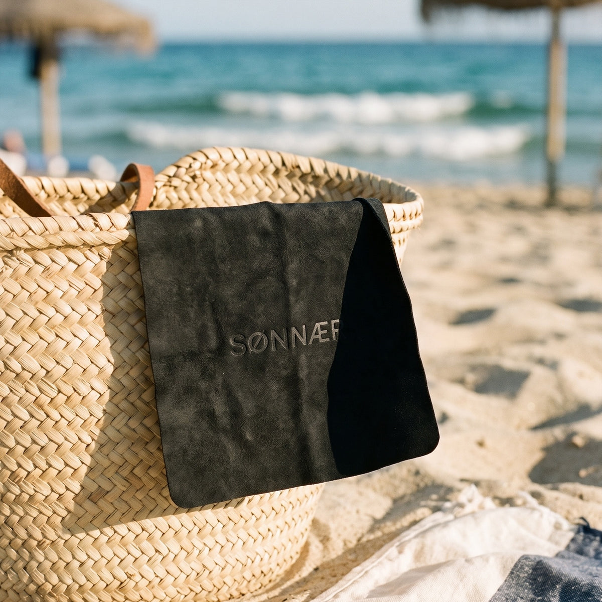 The black suede-finish SØNNÆR cleaning cloth draped over a straw beach bag on the sand