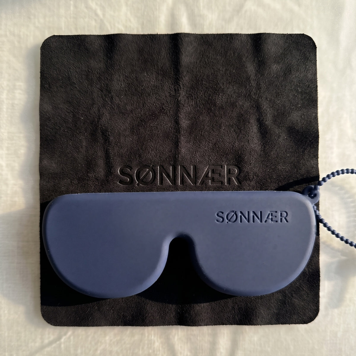 The navy SØNNÆR case resting on its black suede-finish cleaning cloth, with the SØNNÆR mark embossed in the cloth
