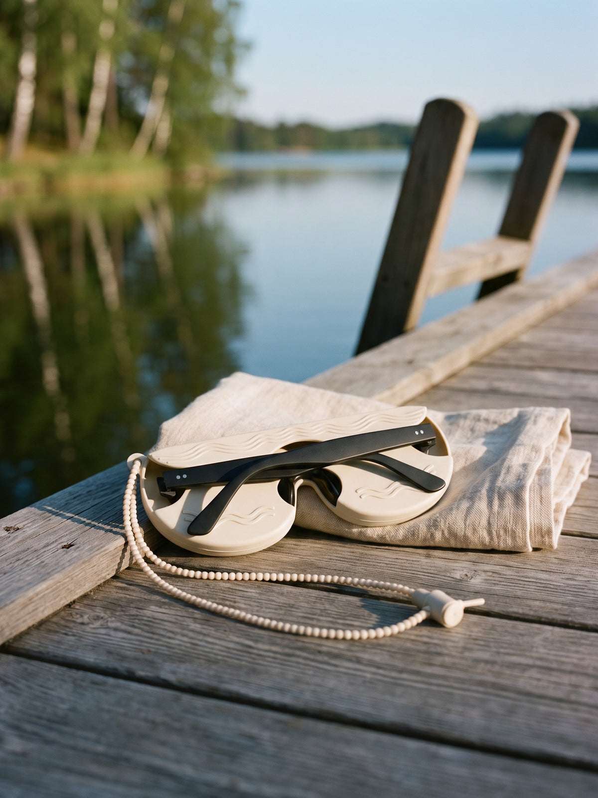 Cream SØNNÆR case holding black sunglasses on a wooden lake jetty