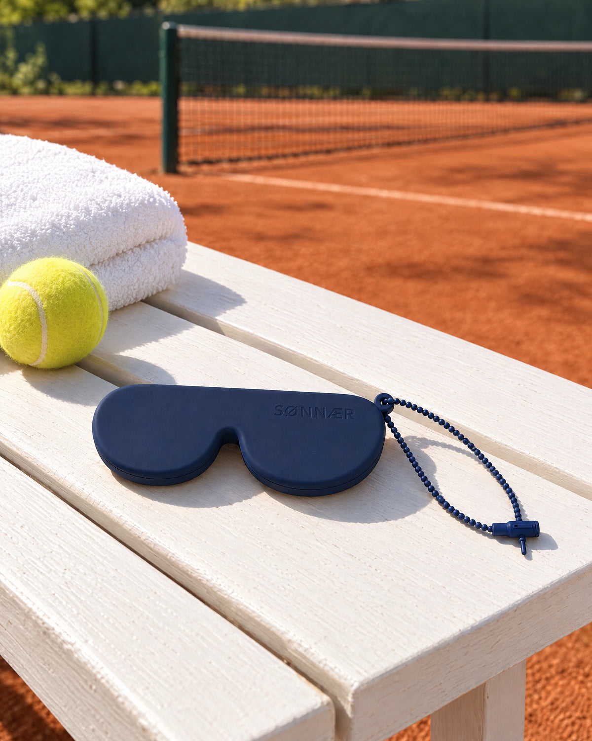 Navy SØNNÆR case on a white bench beside a clay tennis court