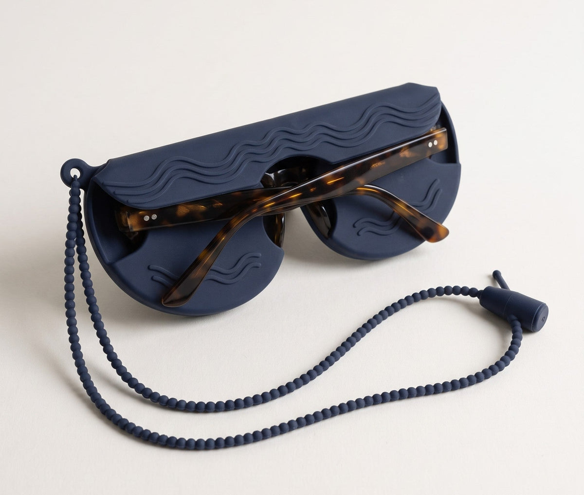 A navy SØNNÆR silicone glasses case, seen from the back, holding a pair of tortoiseshell sunglasses