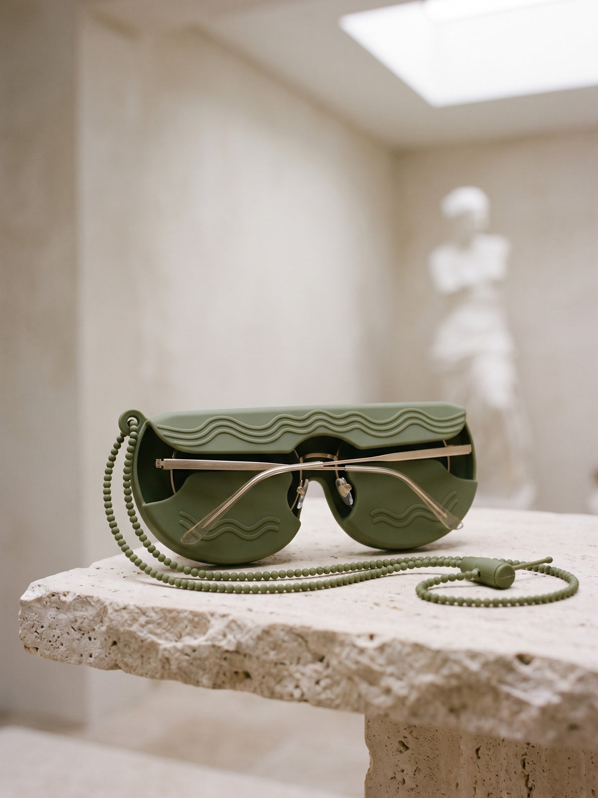 Olive SØNNÆR case holding thin gold-framed sunglasses on a travertine bench in a quiet museum